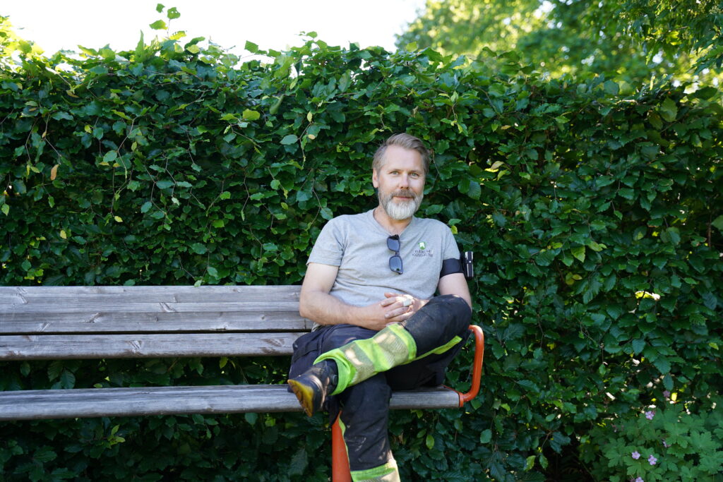 En mann med skjegg sitter på en trebenk foran en grønn hekk. Han har på seg en grå skjorte, svarte bukser med refleksstriper og solbriller som henger på kragen.