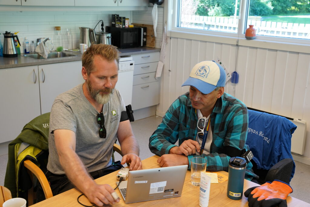 To menn sitter ved et bord på et kjøkken og jobber på en bærbar PC. Det står diverse personlige gjenstander, kopper og flasker på bordet. Miljøet virker uformelt og samarbeidsorientert.