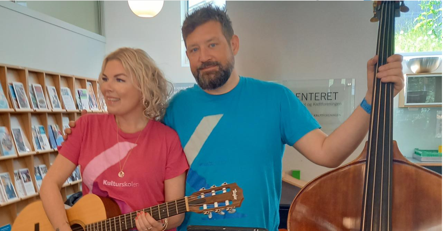 To personer står innendørs; kvinnen holder en akustisk gitar, mens mannen står ved siden av henne med en kontrabass. I bakgrunnen skimtes hyller med bøker og et skilt.