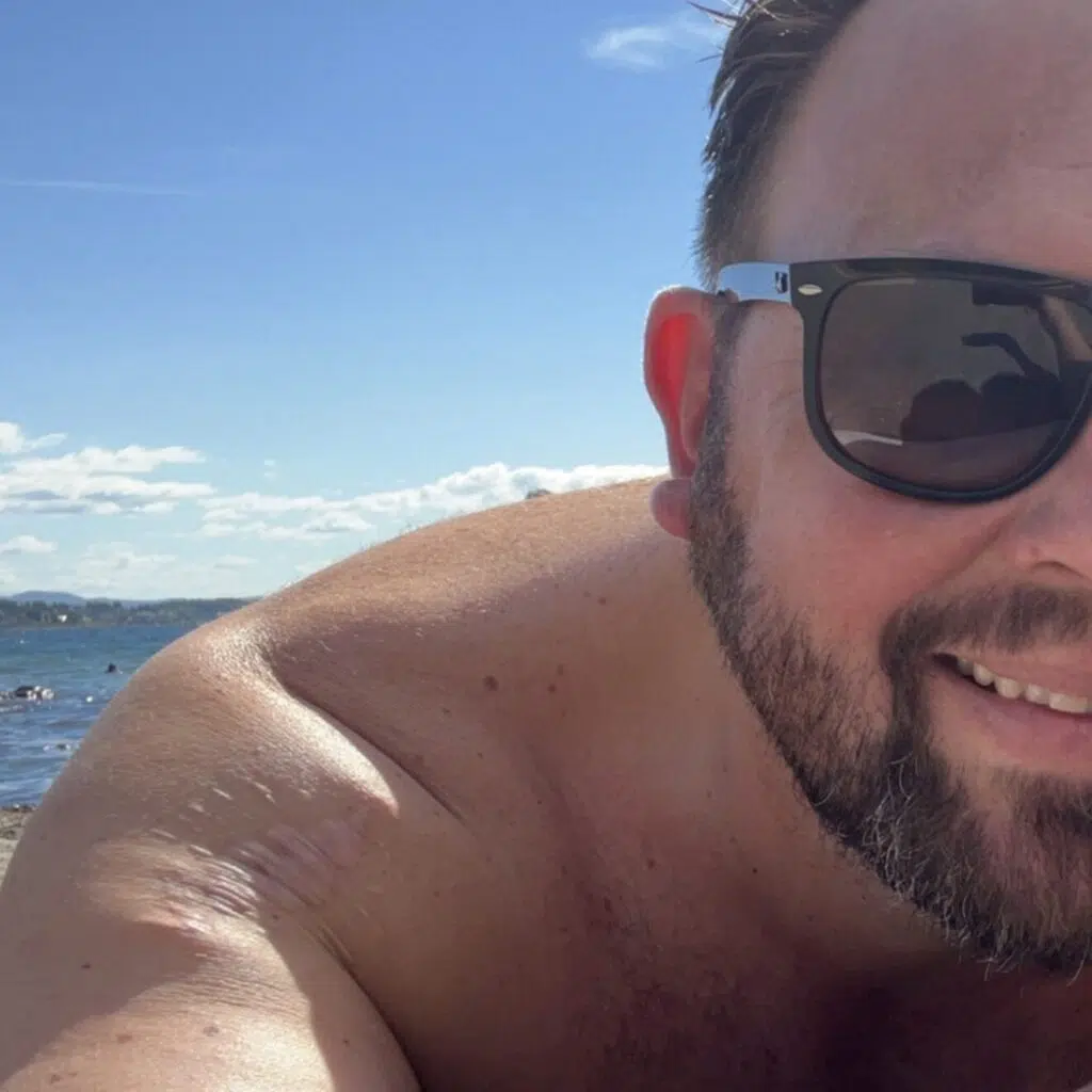 En mann med skjegg og solbriller ligger på en strand under en blå himmel, med vann og fjerne mennesker i bakgrunnen.