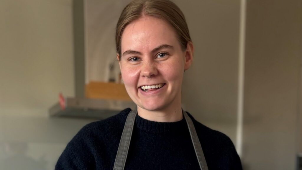 En kvinne med lysebrunt hår, kledd i en mørk genser og et grått forkle, smiler mens hun står inne på et kjøkken.