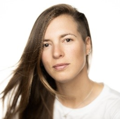 En person med langt brunt hår, delvis barbert på den ene siden, iført hvit overdel, poserer mot en vanlig hvit bakgrunn.