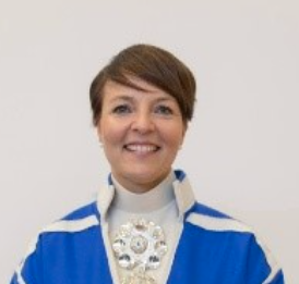 En person med kort, brunt hår, iført blå og hvit seremoniell kappe og en stor, utsmykket sølvmedaljong, står mot en ensfarget, lys bakgrunn.