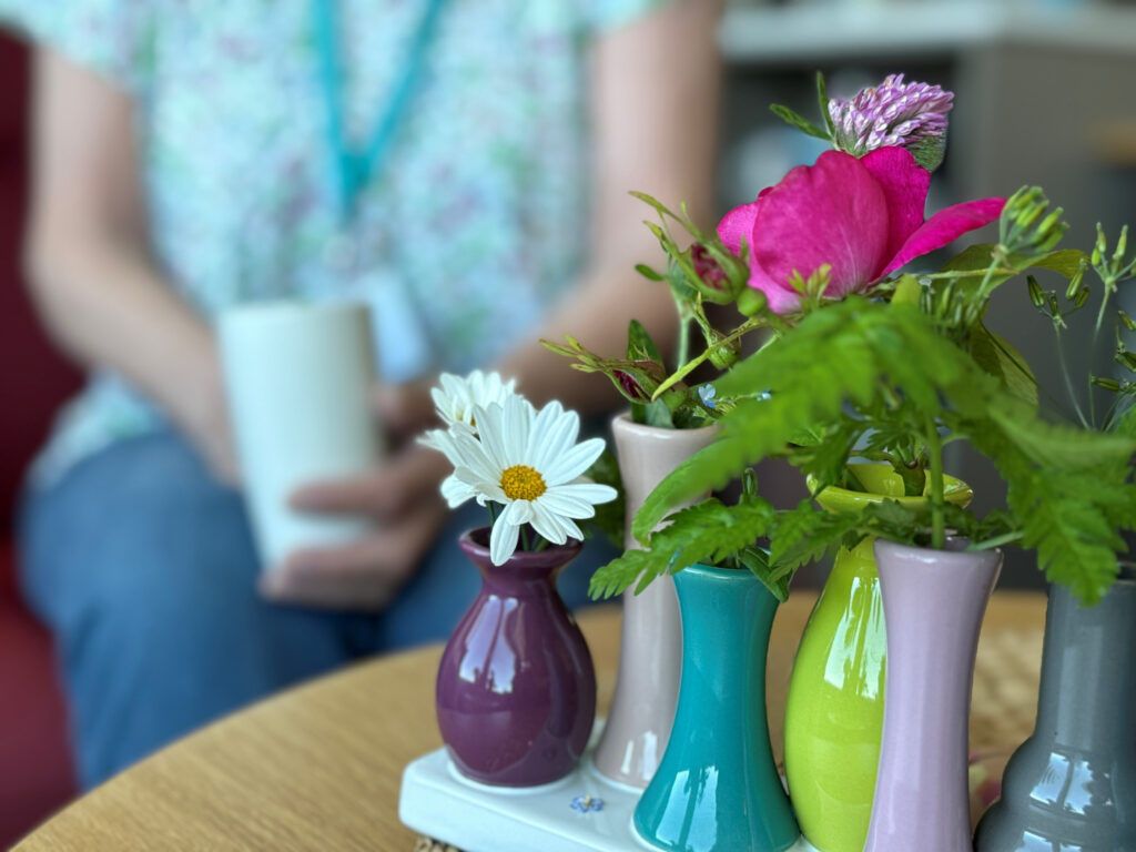 På et bord står en rekke små, fargerike vaser med ulike blomster; i bakgrunnen ser man en uskarp skikkelse som holder en hvit kopp.