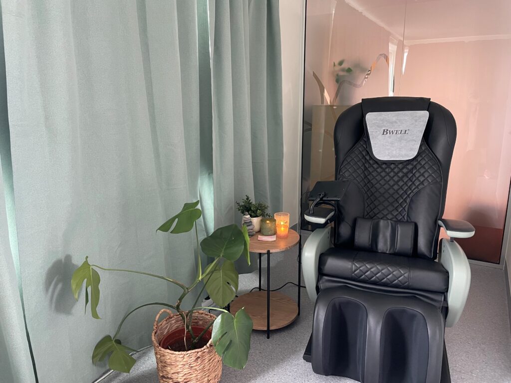 En svart massasjestol står ved siden av et lite rundt bord med stearinlys, en plante og en drink, ved siden av lysegrønne gardiner og en potteplante på gulvet.