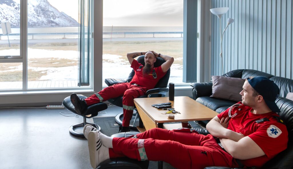 To ambulansepersonell i røde uniformer slapper av i svarte stoler i en salong med et stort vindu som gir utsikt over et snødekt fjell og havet utenfor.