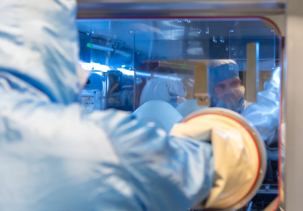To personer i vernedrakter og masker arbeider i et laboratoriemiljø bak glass, og bruker hanskeåpninger for å håndtere materialer i et kontrollert miljø.