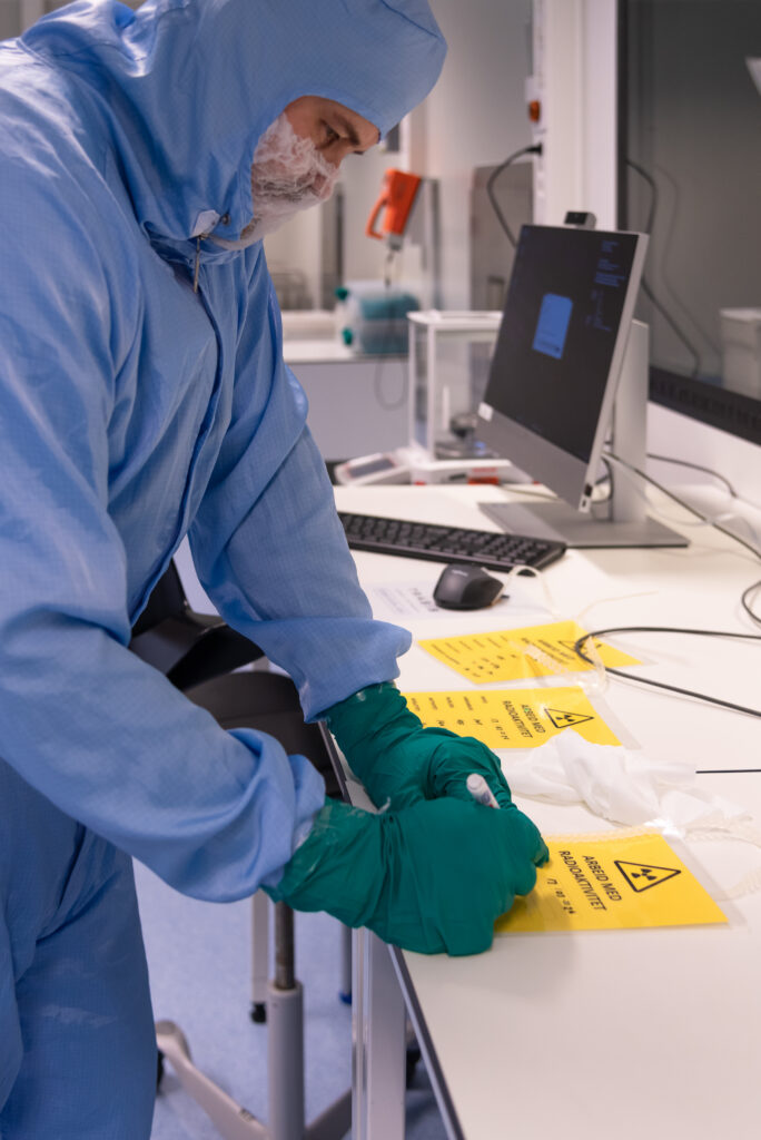 En person i verneklær og hansker håndterer advarselsskilt for farlig materiale ved en laboratoriearbeidsstasjon med en datamaskin.