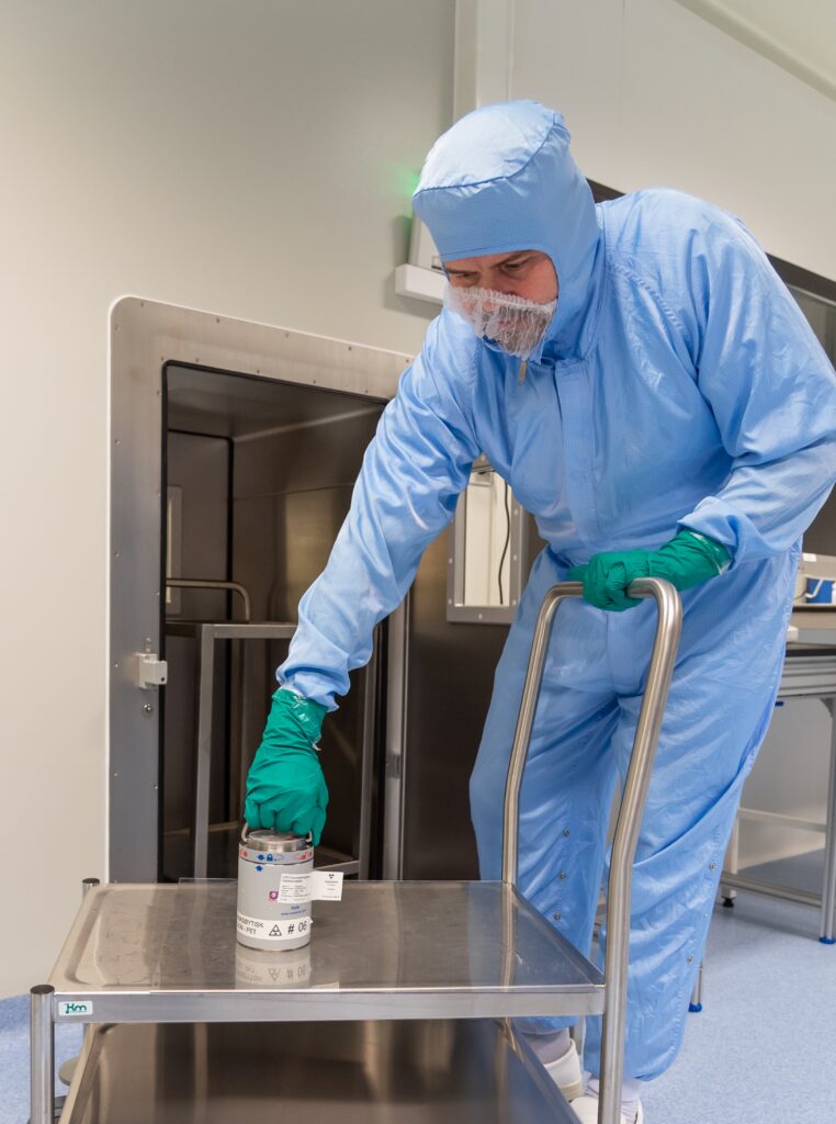 En person i blå verneklær og grønne hansker setter en beholder på en metallvogn i et sterilt laboratoriemiljø.