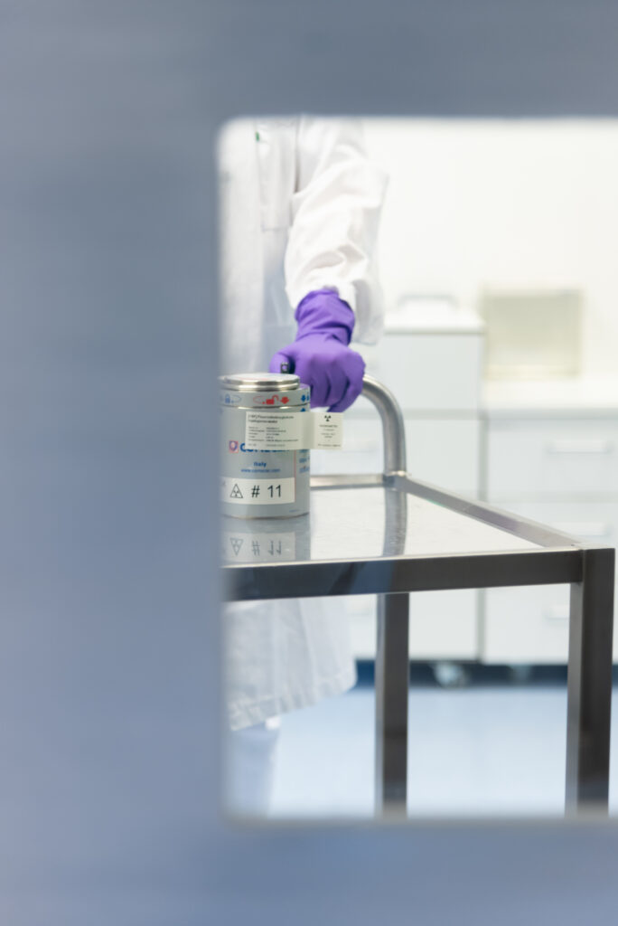 En person i laboratoriefrakk og lilla hansker står ved siden av et metallbord og holder en beholder merket med advarselssymboler i et laboratoriemiljø.