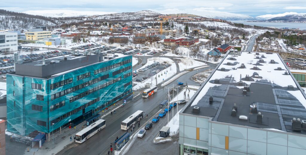 Et snødekt bylandskap med en moderne bygning i blågrønt glass, busser ved en holdeplass, parkerte biler og boligområder på åsene i bakgrunnen.