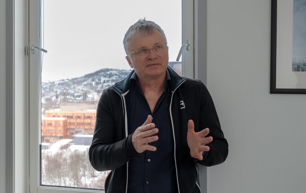 En eldre mann med briller gestikulerer mens han snakker, stående innendørs foran et vindu med utsikt over snødekte bygninger og åser.