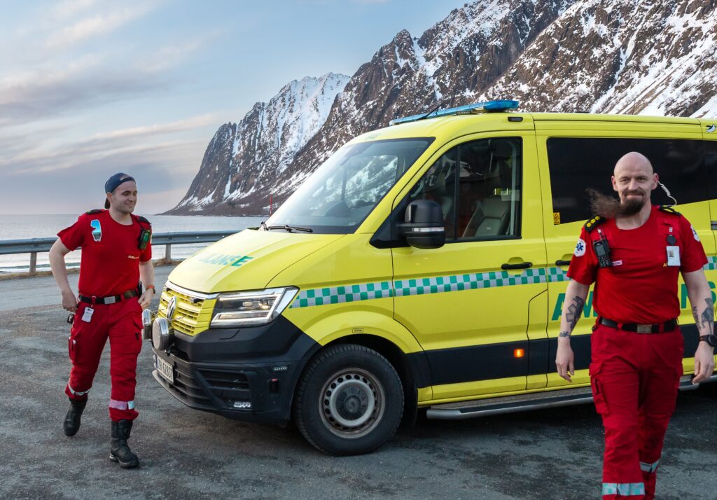 To ambulansepersonell i røde uniformer står ved siden av en gul ambulanse som står parkert utendørs, med snødekte fjell i bakgrunnen.