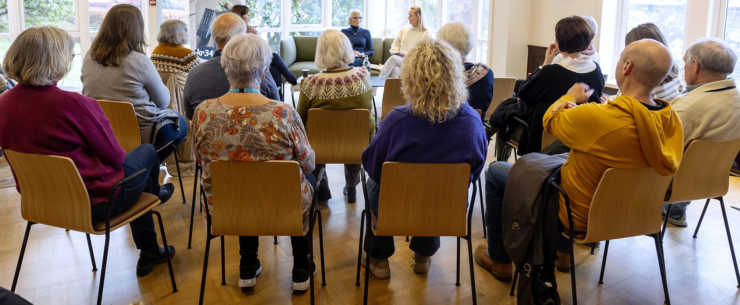 En gruppe voksne sitter på stoler som er plassert i en halvcirkel og lytter til to personer som snakker foran i et lyst rom med store vinduer.