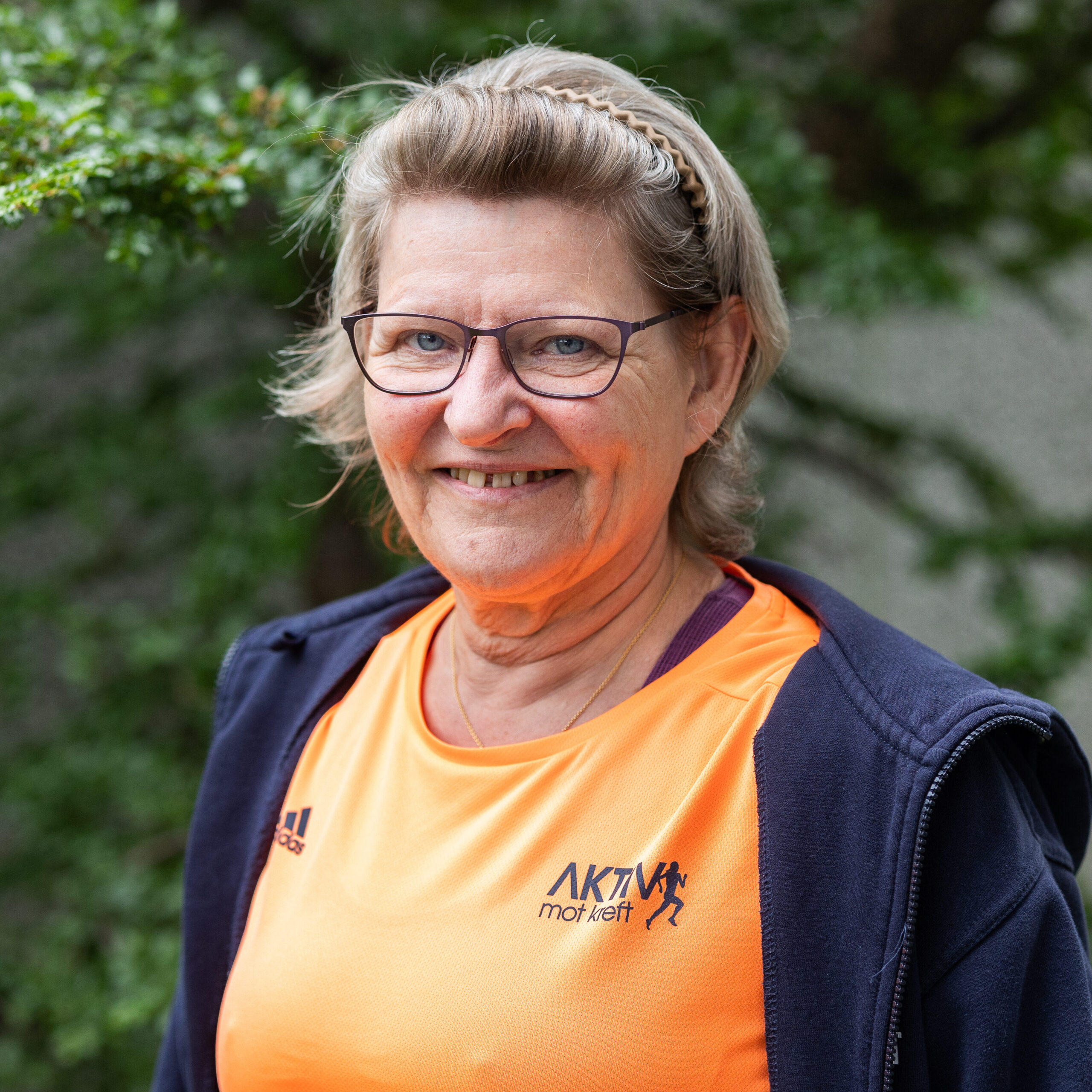 En eldre kvinne med briller og et hårbånd smiler ute, kledd i en oransje sportstrøye og en marineblå jakke.