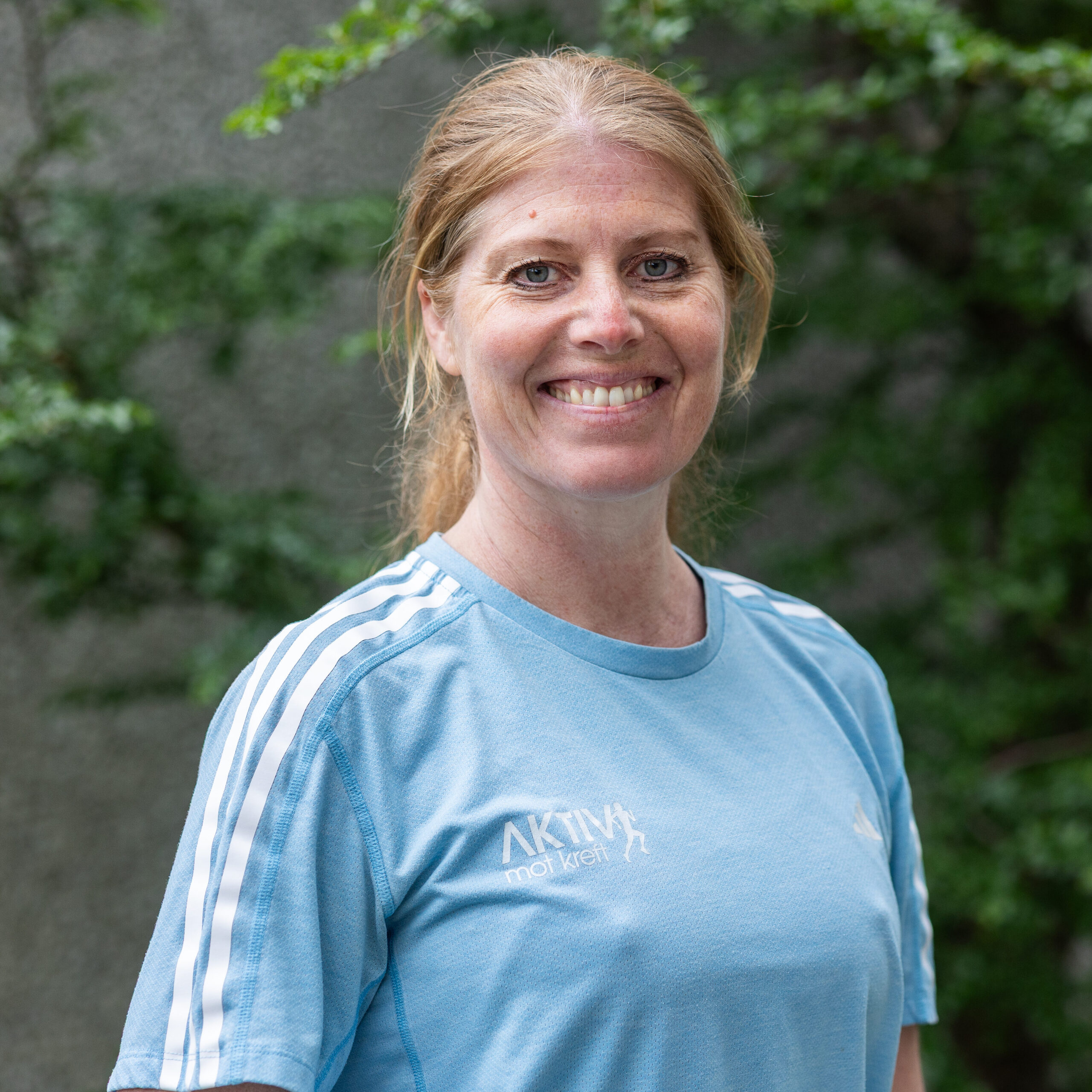 En kvinne med lysebrunt hår som er satt opp, smiler til kameraet. Hun har på seg en lyseblå sportsskjorte og står utendørs med grønt løvverk i bakgrunnen.