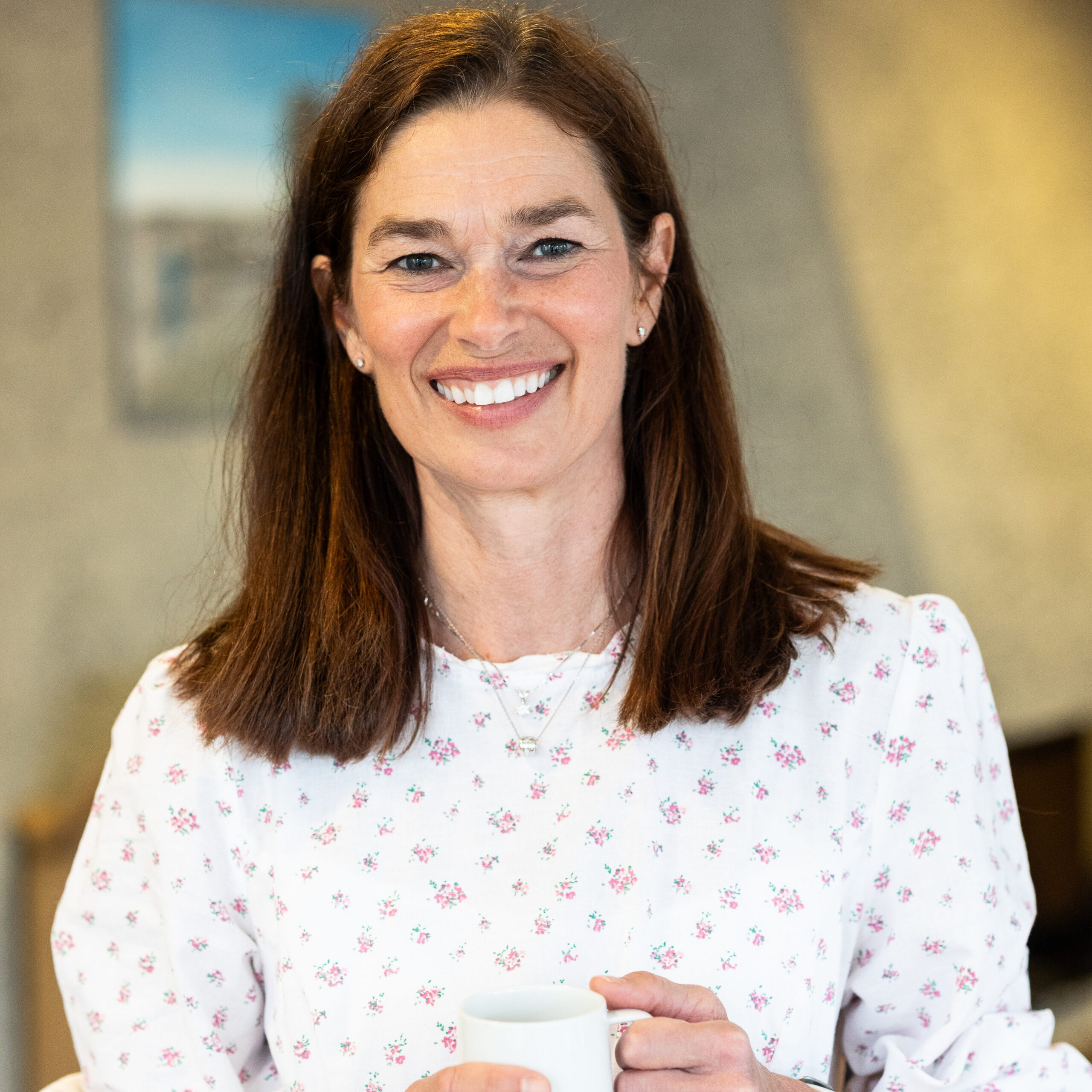 En kvinne med langt brunt hår, kledd i en lys blomstret kjole, står inne og holder en hvit kopp mens hun smiler til kameraet.