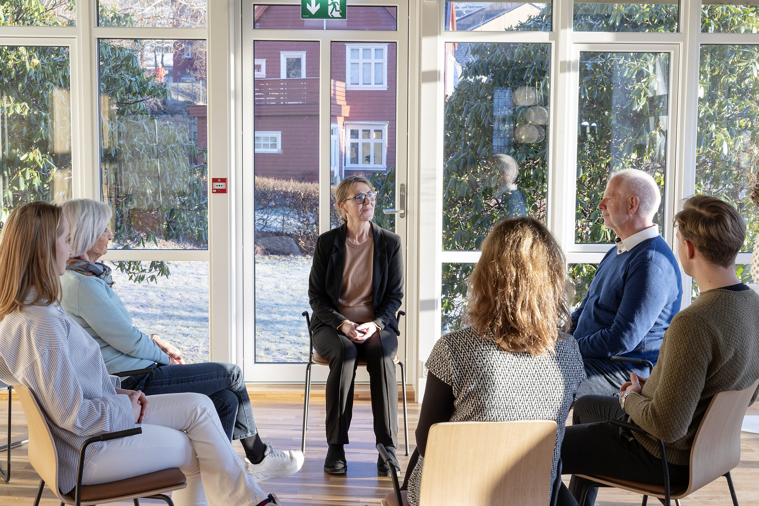 Seks voksne sitter i en halvcirkel innendørs og deltar i en gruppesamtale ved store vinduer der sollyset strømmer inn.
