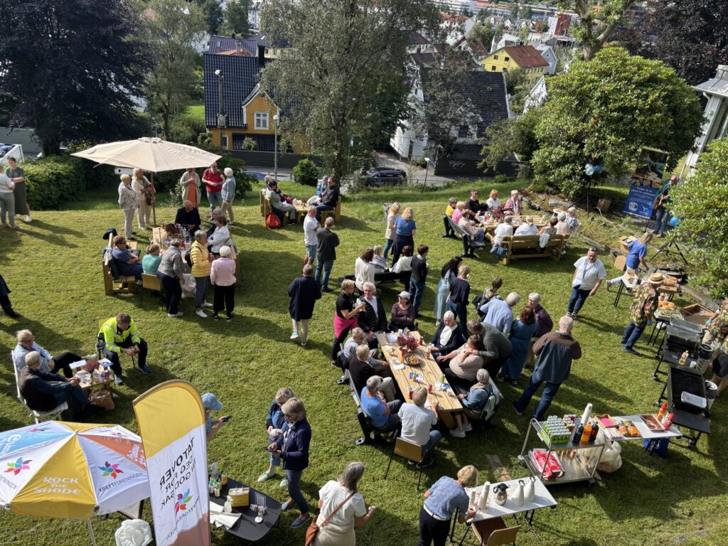 En gruppe mennesker har samlet seg utendørs på en gressplen; de sitter ved bord og står i små grupper, med mat og drikke i bildet og hus i bakgrunnen.