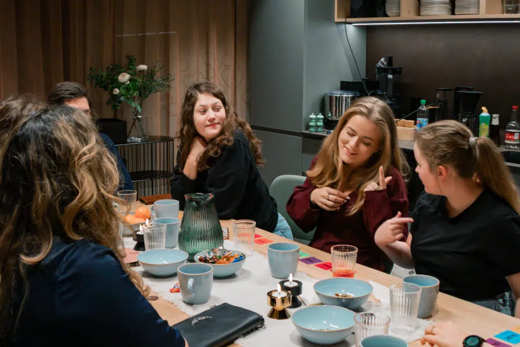 En gruppe mennesker sitter rundt et bord med skåler, glass og snacks, og snakker sammen i et rom med varm belysning.