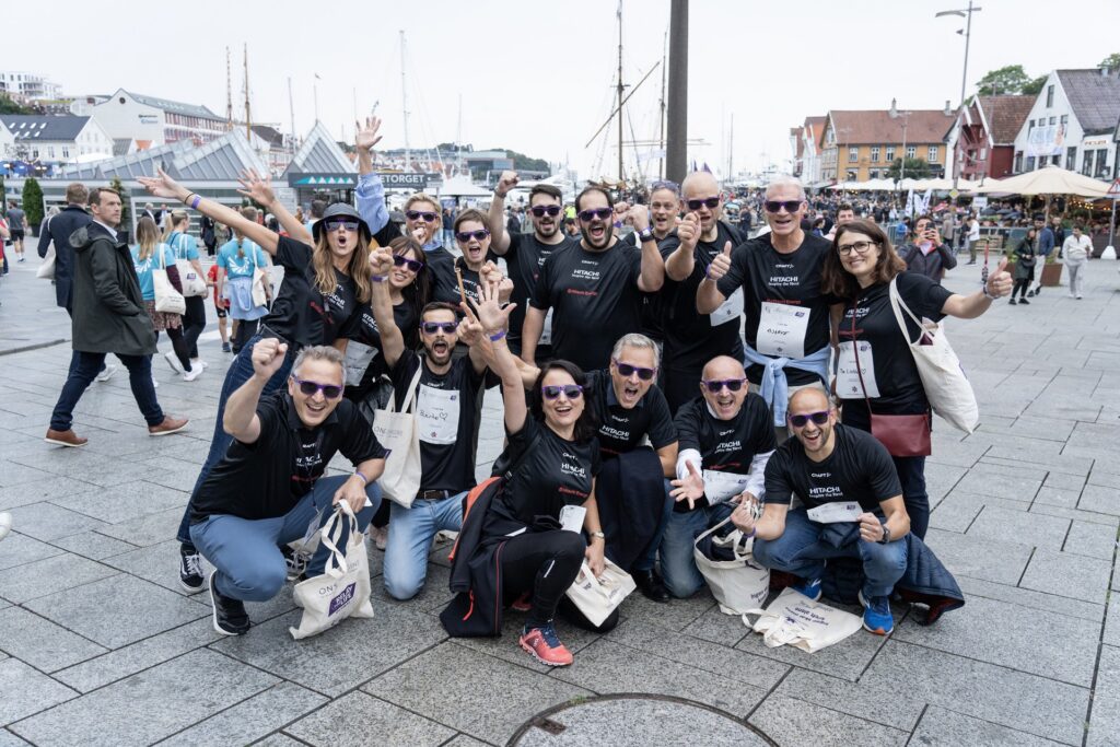 En gruppe mennesker kledd i matchende svarte skjorter og solbriller poserer sammen utendørs i et livlig byområde med bygninger og markedsboder i bakgrunnen.
