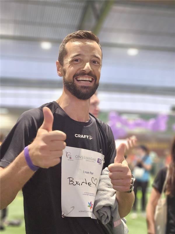 En mann innendørs under et arrangement gir to tommeler opp og smiler. Han har på seg et startnummer der det står «I run for Barde♥». Folk og idrettshallen i bakgrunnen er synlige, men uskarpe.