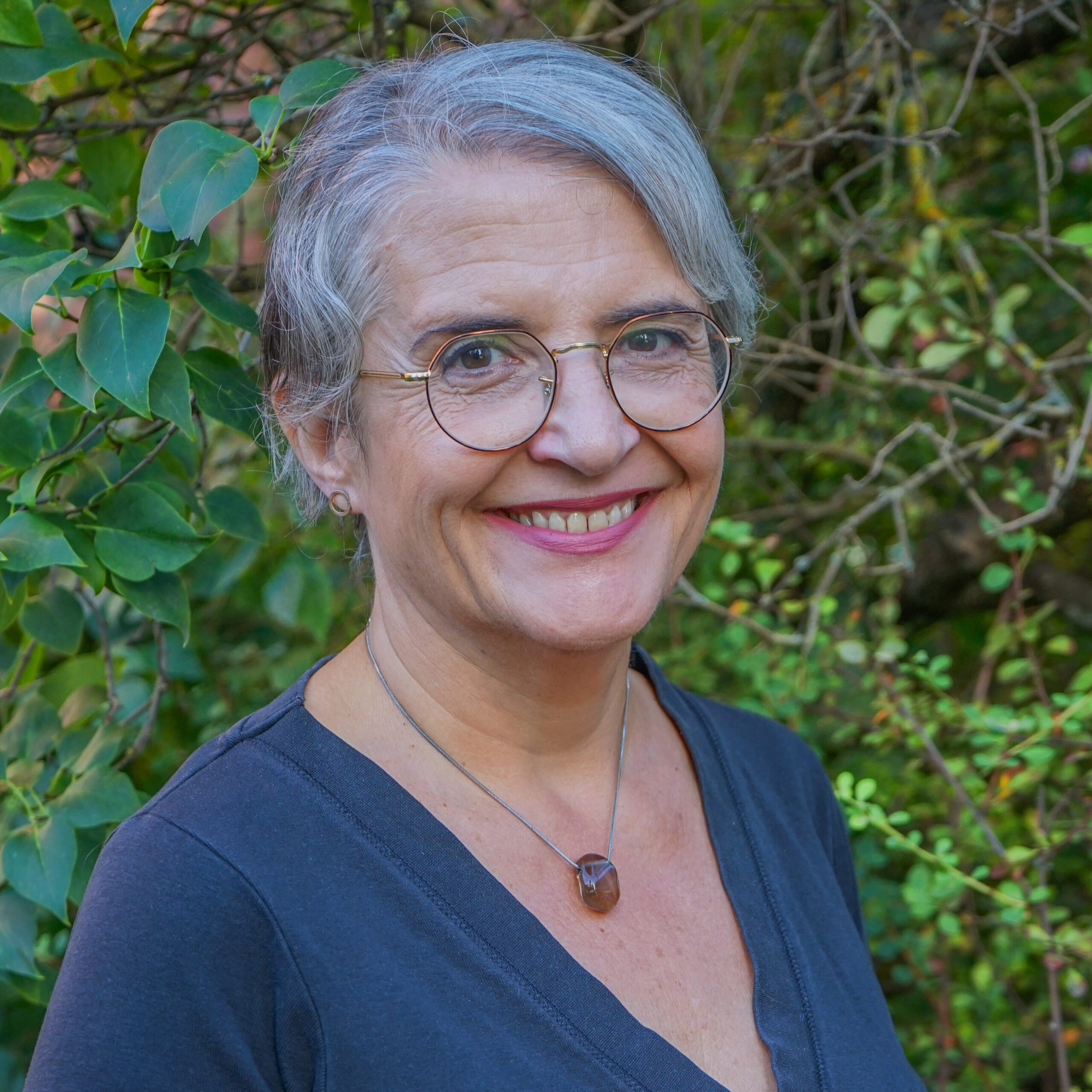 En eldre kvinne med kort, grått hår og briller, kledd i en mørk topp og et halskjede med steinanheng, som står utendørs foran grønt løvverk og smiler til kameraet.