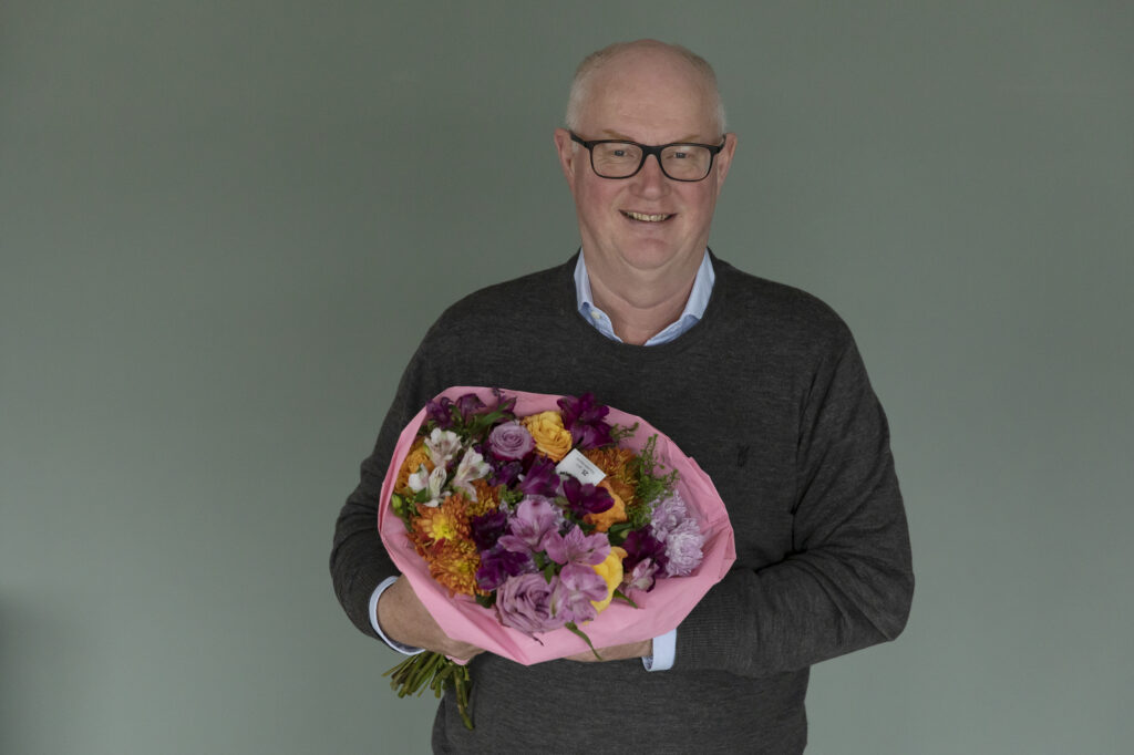 En mann med briller og en mørk genser står foran en ensfarget bakgrunn og holder en bukett med fargerike blomster innpakket i rosa papir.