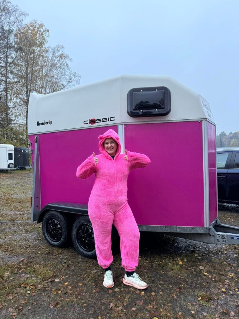 En person kledd i en knallrosa, lodden drakt står foran en rosa og hvit campingvogn og strekker begge tommelfingrene opp på en overskyet dag, med høsttrær i bakgrunnen.