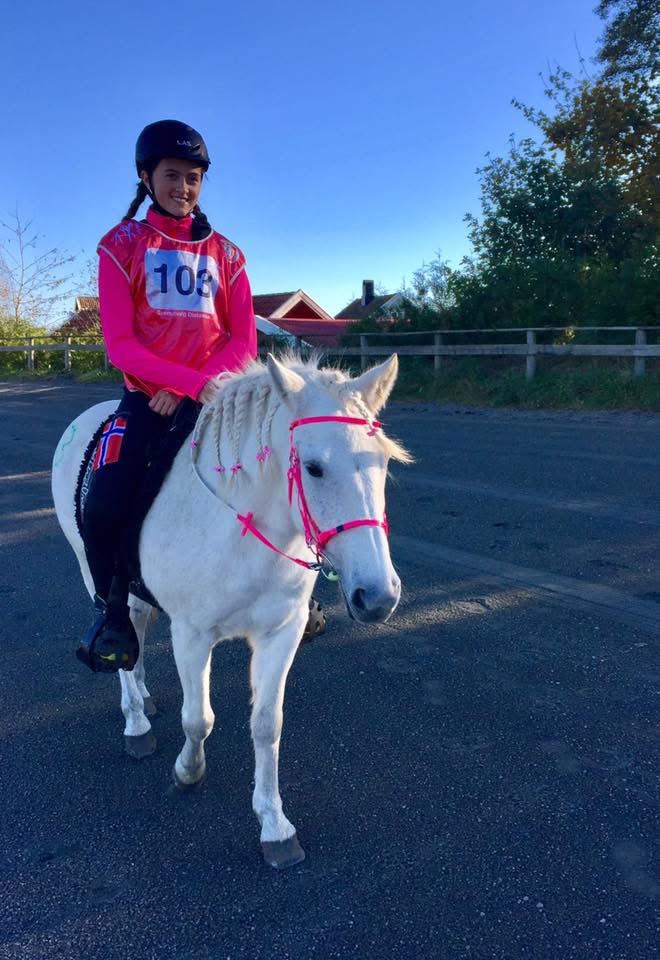 En person med hjelm og startnummer rir på en hvit hest med rosa rideutstyr på en asfaltert vei, med hus og trær i bakgrunnen under en klar blå himmel.
