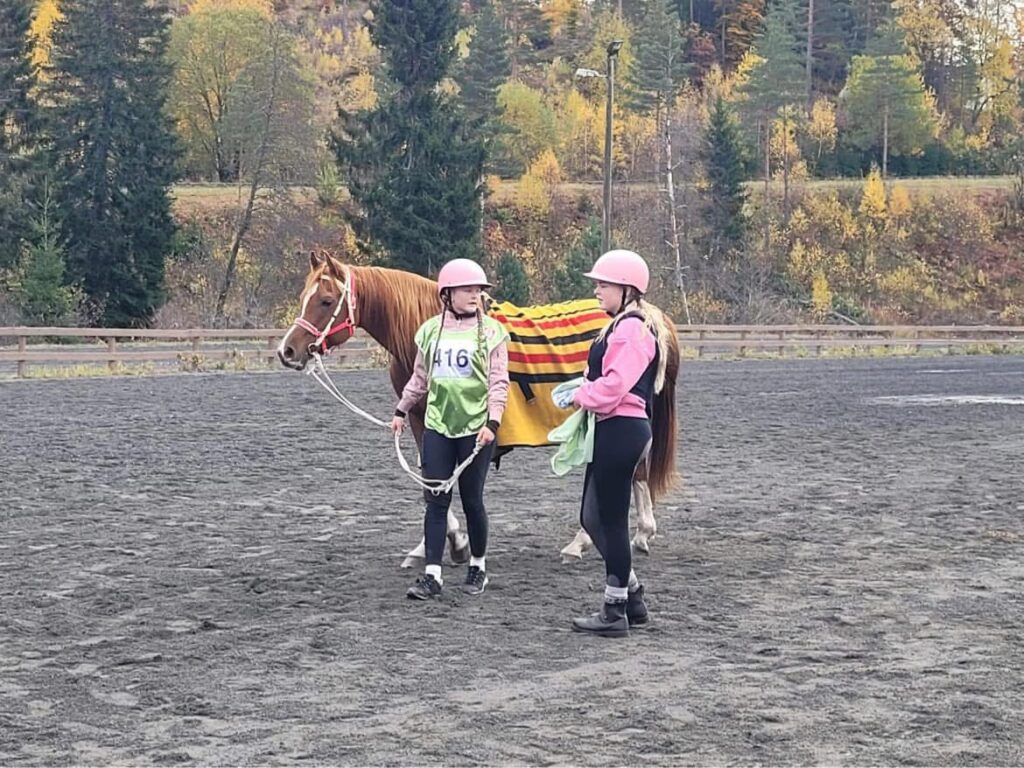 To personer med hjelmer og nummererte vester står sammen med en hest som er dekket av et stripete teppe på en grusbane med trær i bakgrunnen.