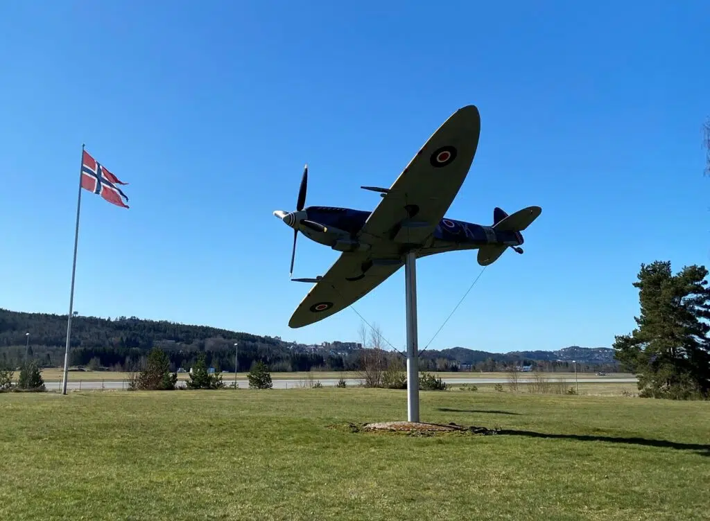 Et Spitfire-jagerfly står oppstilt på en sokkel i et gressområde, med et norsk flagg på en flaggstang og en klar blå himmel i bakgrunnen.