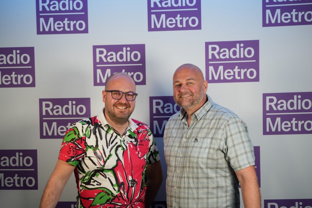To menn står og smiler foran en bakgrunn med flere «Radio Metro»-logoer i lilla og hvitt.