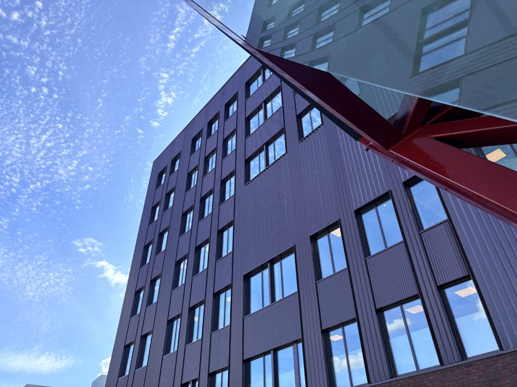 En høy, moderne bygning med mange vinduer sett nedenfra, med en rød arkitektonisk struktur og bygningens speilbilde i glasset, under en blå himmel med spredte skyer.