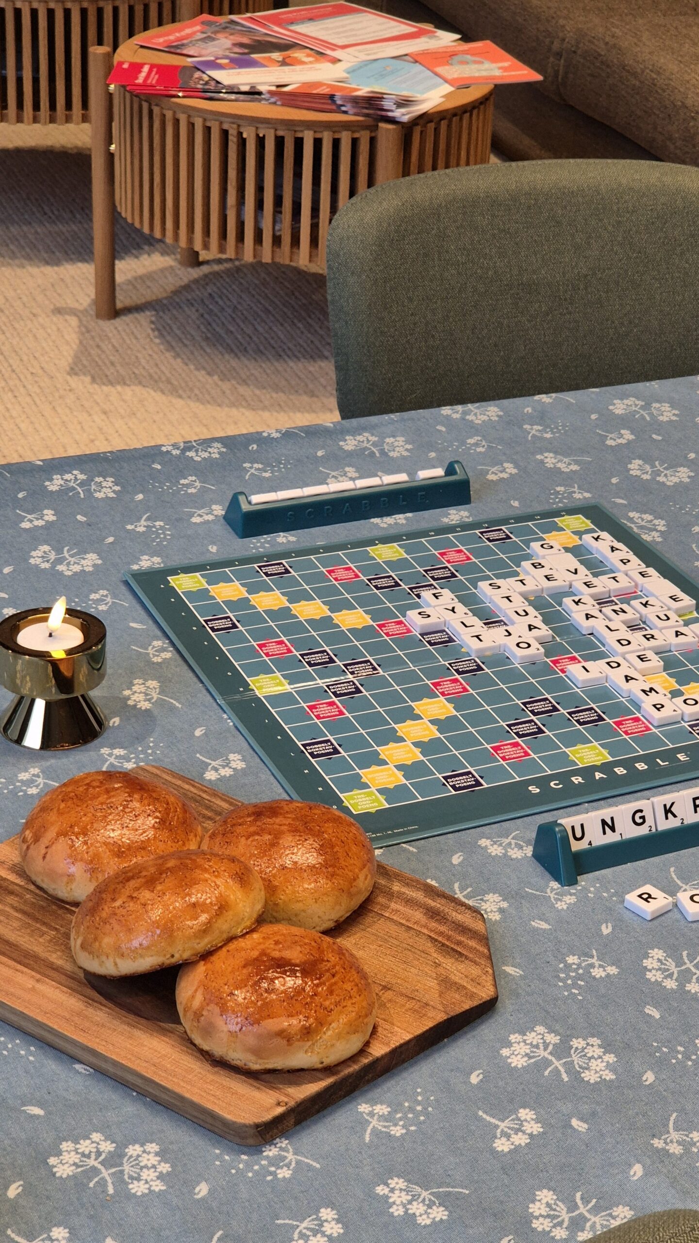 Et Scrabble-spill som er i gang, er plassert på en blå blomstret duk, ved siden av et tent stearinlys, fire boller på et trebrett og blader på et trebord i bakgrunnen.