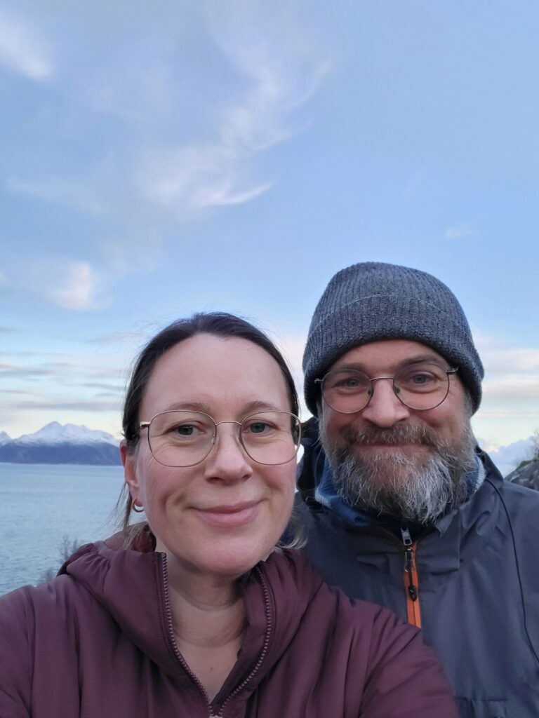 To personer med briller og vinterjakker smiler til kameraet utendørs, med et vann og snødekte fjell i bakgrunnen under en overskyet himmel.