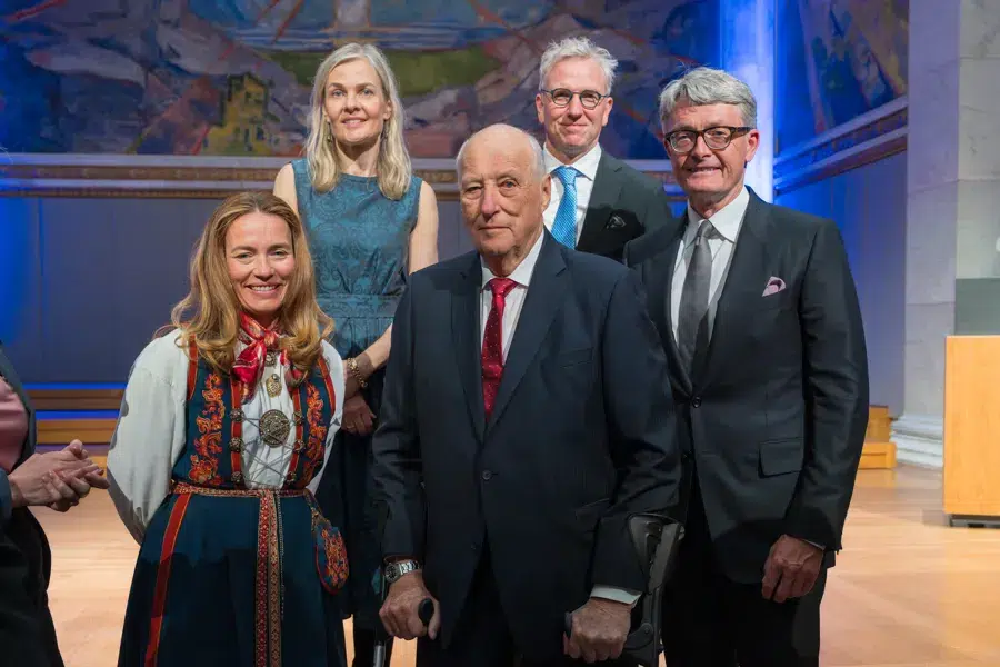 Fem voksne – tre menn i dress og to kvinner i kjoler – poserer sammen innendørs i en formell setting, med kunstverk synlige i bakgrunnen.