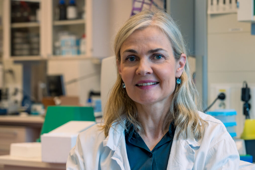 En kvinne i hvit laboratoriefrakk sitter med armene i kors i et laboratorium, med vitenskapelig utstyr i bakgrunnen.