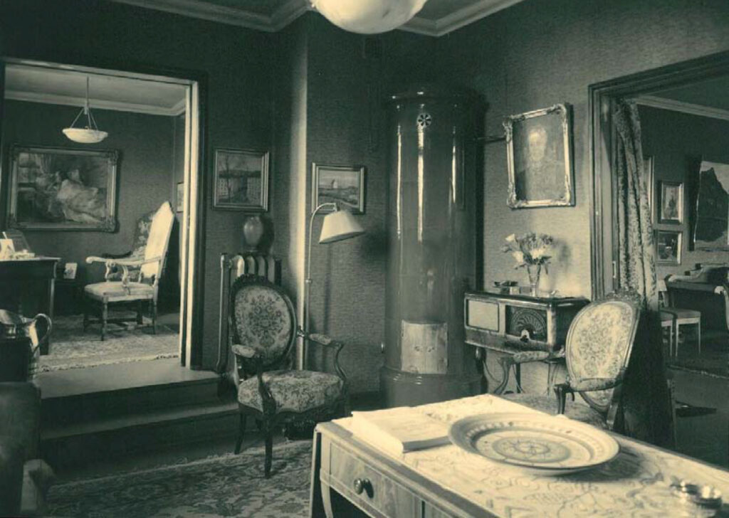 En stue i vintage-stil med utsmykkede stoler, en høy, sylindrisk ovn, innrammede kunstverk, en radio, en lampe og en dekorativ tallerken på et skrivebord; gjennom en åpen døråpning kan man se et annet rom.
