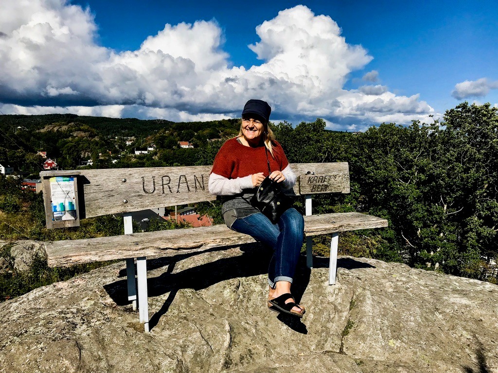 En person i genser og lue sitter på en trebenk på toppen av en steinete ås, med trær, skyer og hus i bakgrunnen.