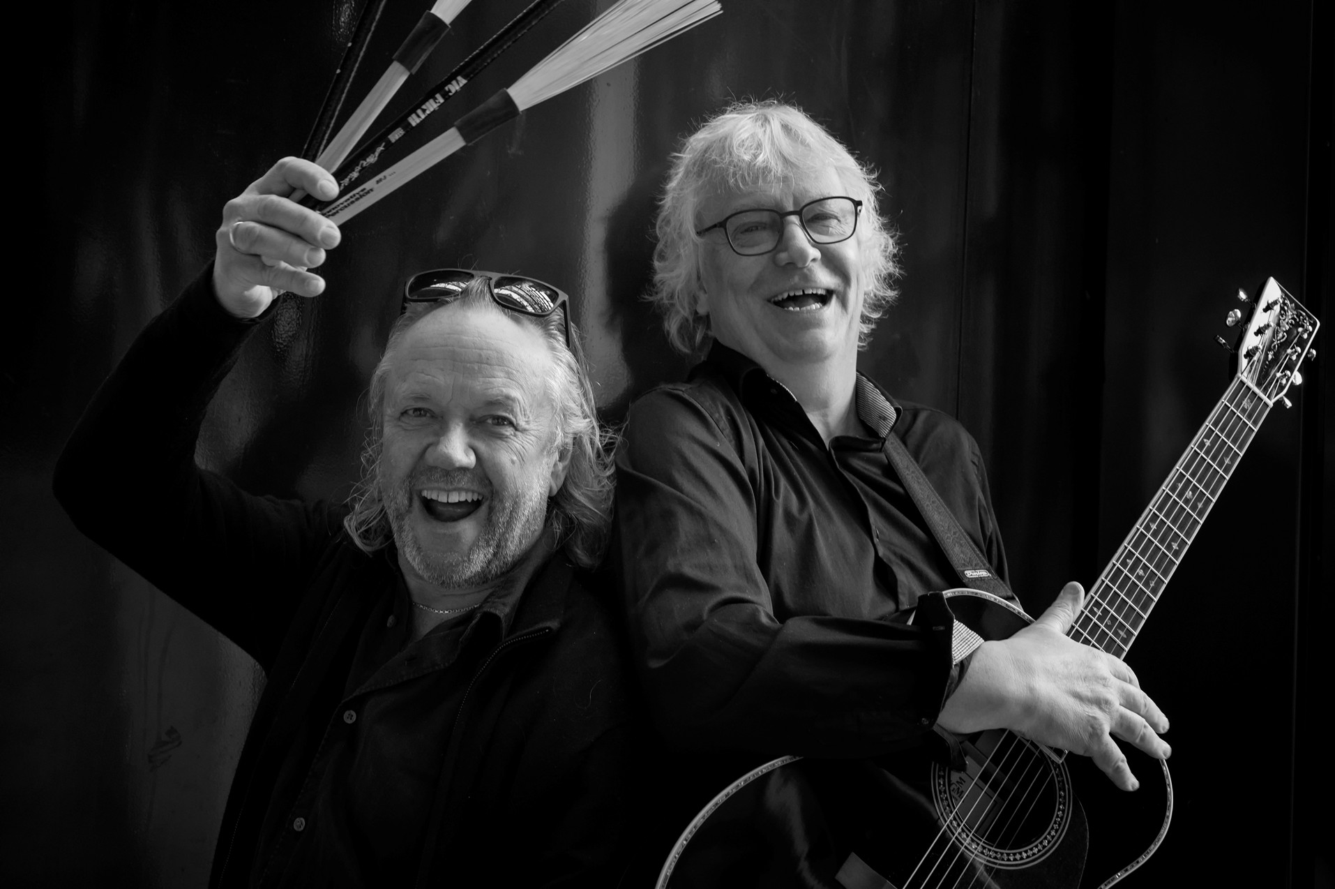 To eldre menn som smiler; den ene holder trommestikker over hodet, den andre holder en akustisk gitar. Begge har på seg mørke skjorter og har langt, lyst hår. Bakgrunnen er ensfarget og mørk.