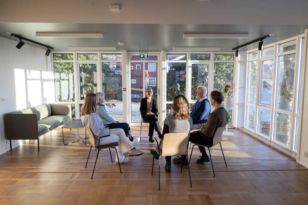 Seks personer sitter i en sirkel og diskuterer i et lyst rom med store vinduer, tregulv og moderne møbler.