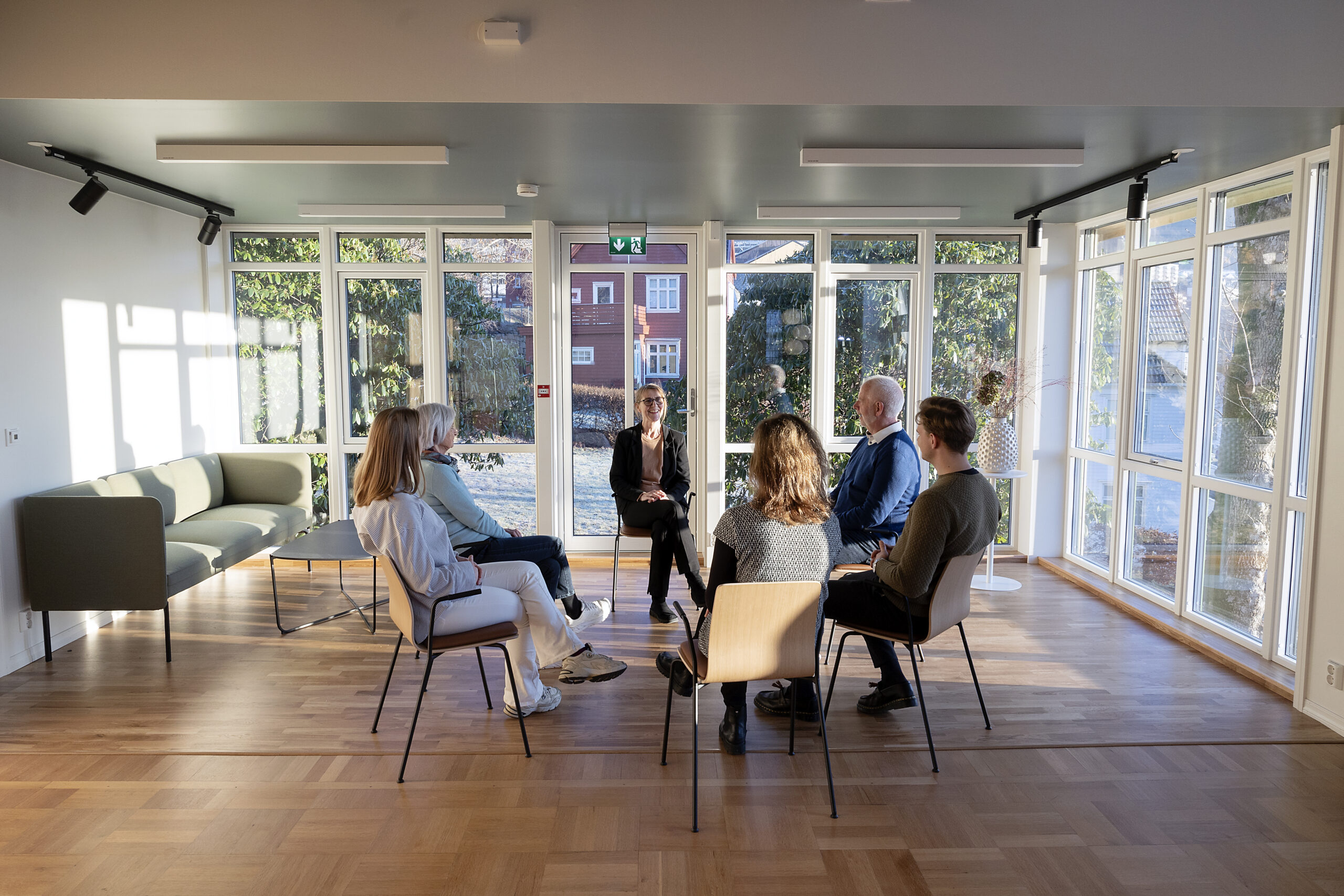 Seks personer sitter i en sirkel og diskuterer i et lyst rom med store vinduer, tregulv og moderne møbler.