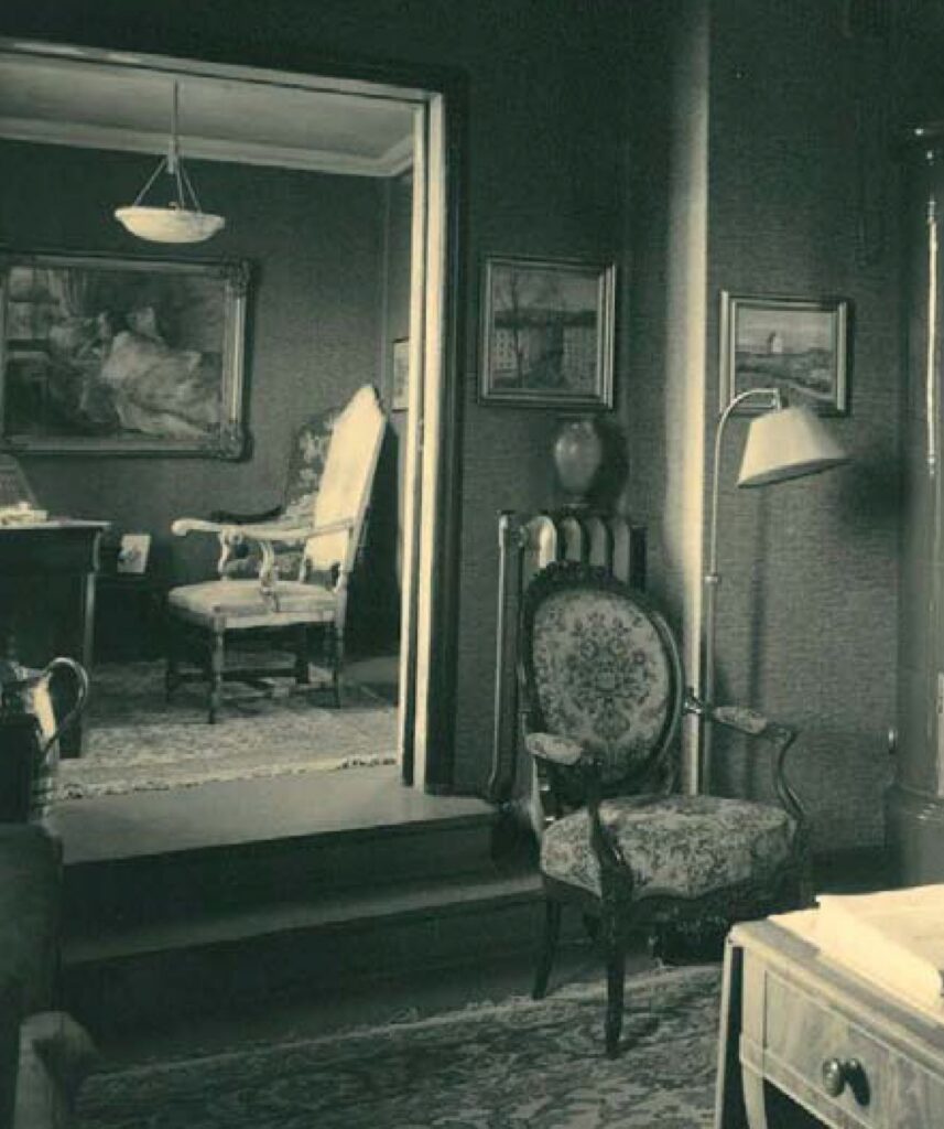 En stue i vintage-stil med utsmykkede stoler, innrammede malerier på veggene, et skrivebord og en gulvlampe. En døråpning fører til et annet rom med lignende innredning.
