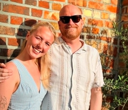 To voksne står side om side foran en murvegg og smiler til kameraet. Personen til venstre har på seg en lyseblå kjole; personen til høyre har på seg solbriller og en lys skjorte.