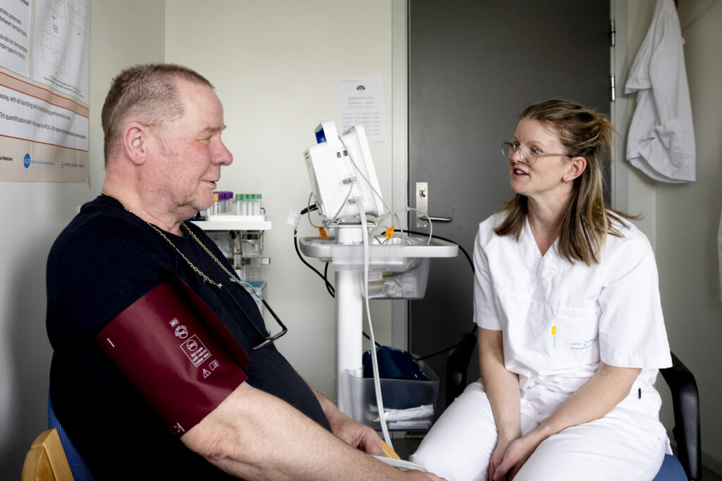 En helsearbeider i hvit uniform snakker med en pasient som sitter med en blodtrykksmansjett på seg i et undersøkelsesrom.
