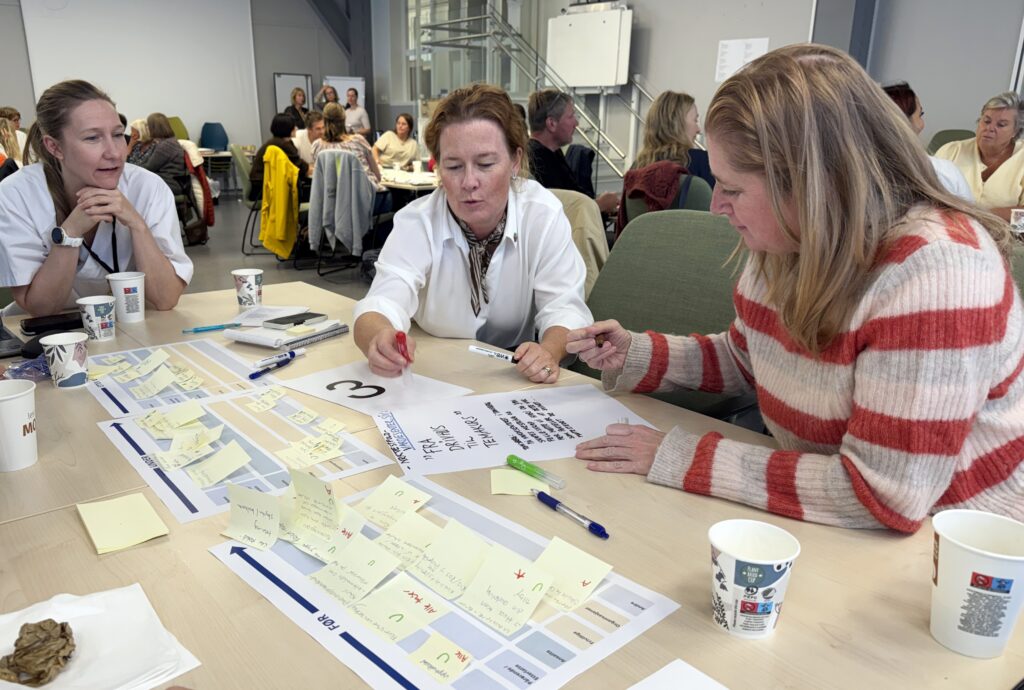 Tre kvinner samarbeider ved et bord dekket av post-it-lapper, tusjer og papirer under et seminar i et travlt, moderne møterom.