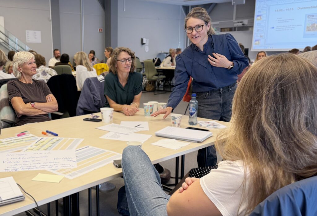 En gruppe mennesker sitter rundt et bord dekket av papirer, mens en kvinne står og gestikulerer og involverer de andre i en diskusjon under et seminar eller et møte.