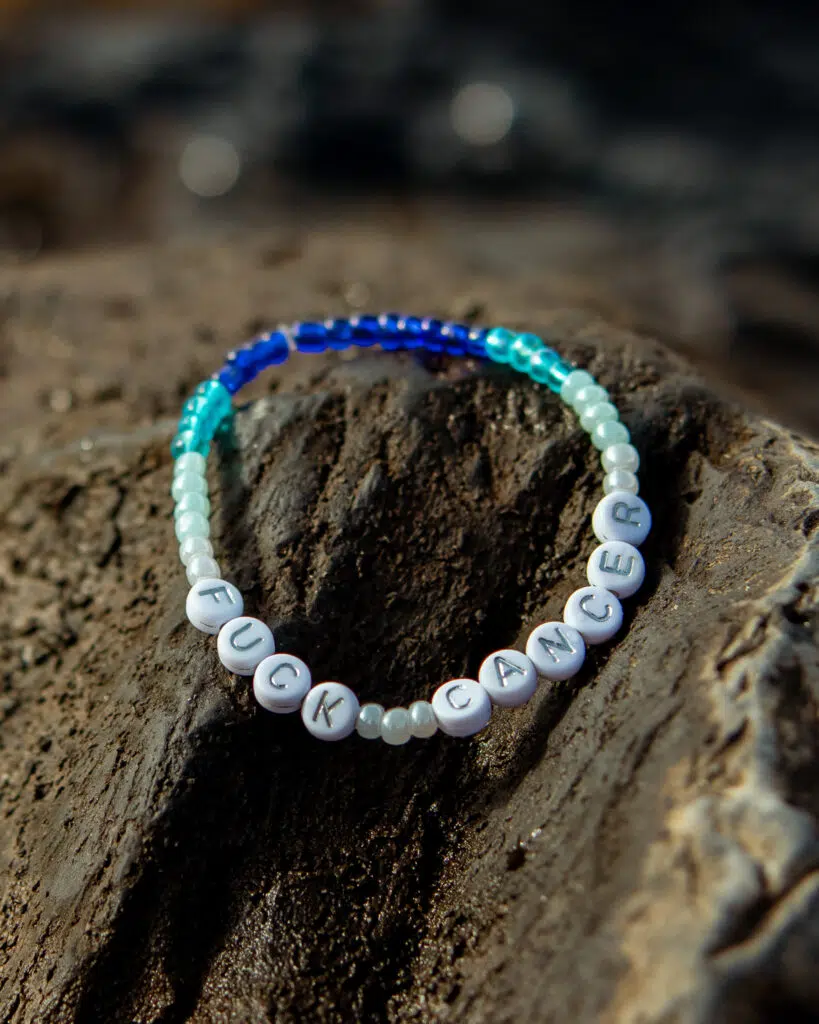 Perlearmbånd på en stein, med hvite perler som danner ordet «FUCK CANCER» og i tillegg blå og turkise perler.