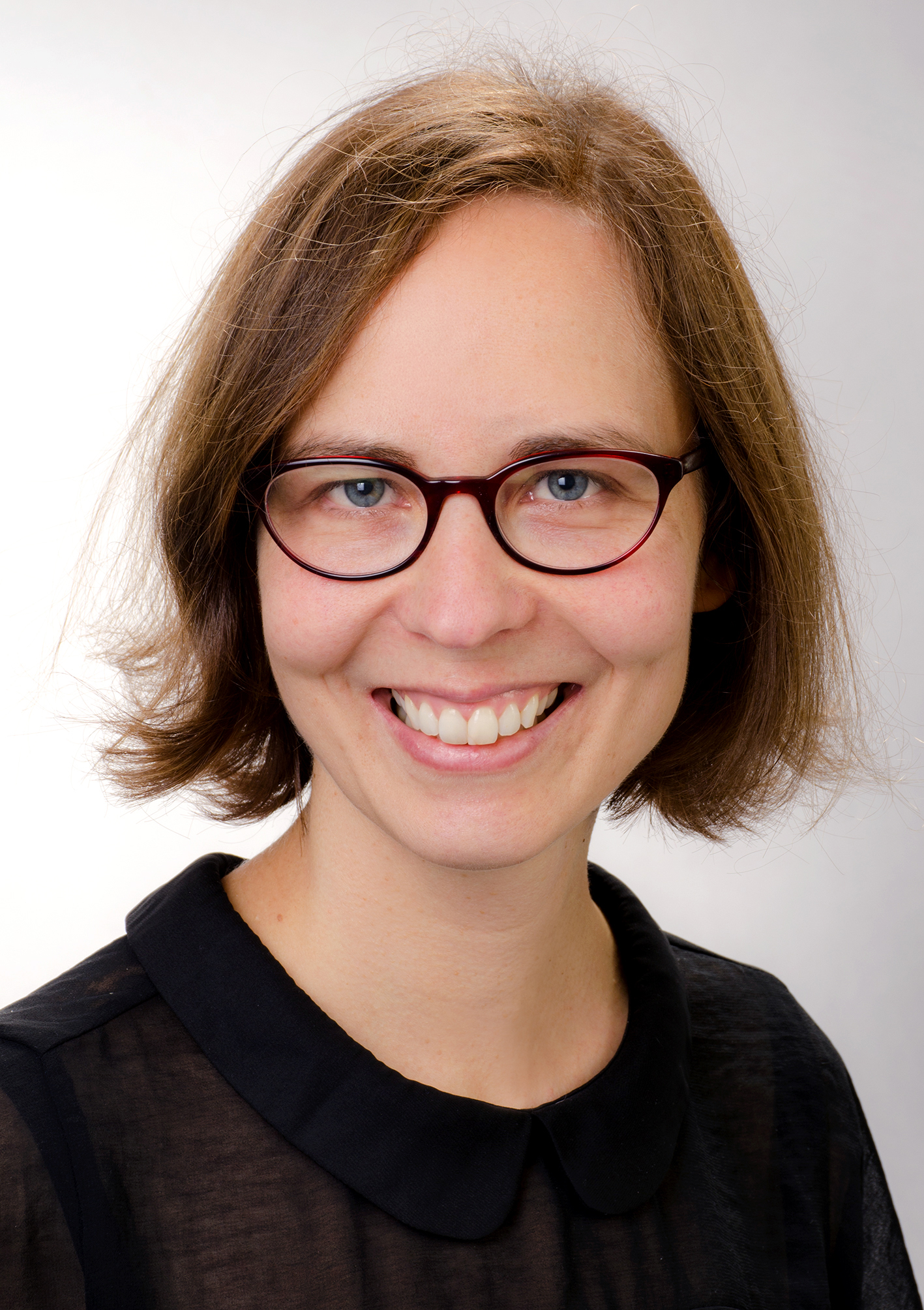 En kvinne med kort brunt hår og briller, kledd i en svart skjorte med krage, som smiler til kameraet mot en ensfarget, lys bakgrunn.